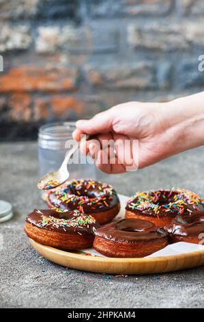 Fried doughnuts with chocolate glaze. Woman's hand decorating doughnuts with colorful sprinkles. Stock Photo