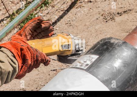 The hands of an industrial worker in working mittens use an electric-grinding equipment tool on a polyethylene water pipe on a construction site. Stock Photo
