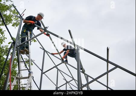Man in safety helmet standing on ladder and holding hammer while colleague installing metal rail for solar modules. Male workers assembling support structures for solar panels under white sky. Stock Photo