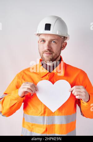 Construction Day concept. Portrait of bearded male construction worker, supervisor or foreman standing by the wall wearing white hard hat and orange reflective vest holding white paper heart shape Stock Photo