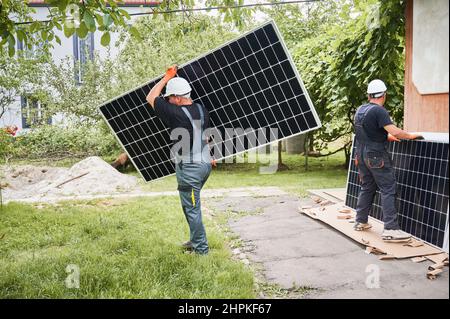 Full length of two men in safety helmets carrying solar modules in the yard. Male worker holding photovoltaic solar panel and walking on grass while colleague standing by house with solar module. Stock Photo