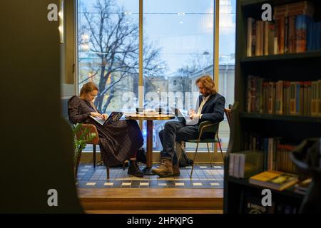 Bored woman and serious focused man sitting together at table in cafe by window working on laptops Stock Photo