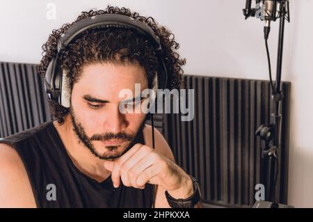 closeup portrait of caucasian man with curlers and with headphones in his home studio looking down touching his chin, concentrating and thinking while Stock Photo