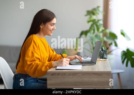 Positive young woman writing in notebook during online lecture on laptop, communicating to colleagues on webcam Stock Photo