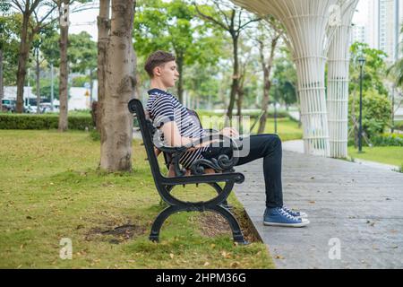 Young caucasian man sitting on the bench in the park and working on the laptop. Student typing on computer, freelance, working online. High quality Stock Photo