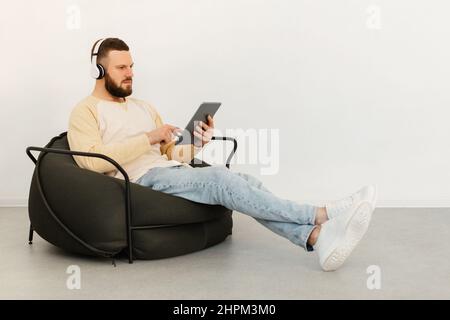 Serious Guy Using Tablet Wearing Earphones Sitting Indoor, Side View Stock Photo