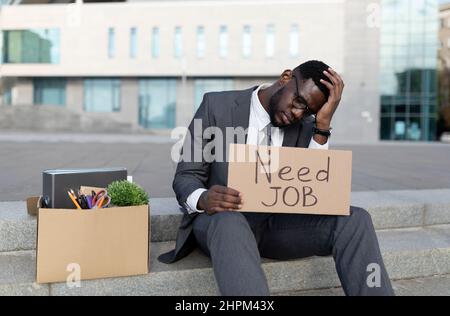 Unhappy black businessman sitting outdoors with box of office supplies and poster with need job text Stock Photo