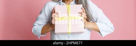 Emotional Asian woman with loose grey hair and party hat holds present on pink background Stock Photo