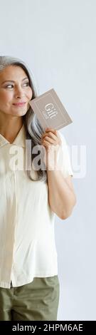 Thoughtful hoary haired Asian lady customer holds gift card standing on light grey background Stock Photo