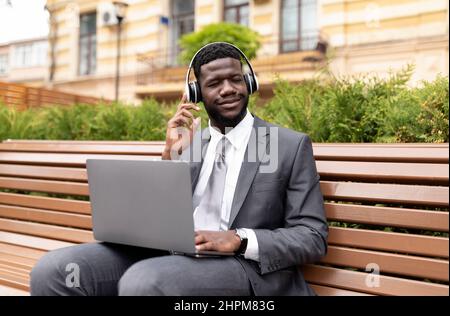 African american businessman in wireless headphones using laptop and listening music with closed eyes, sitting outdoors Stock Photo