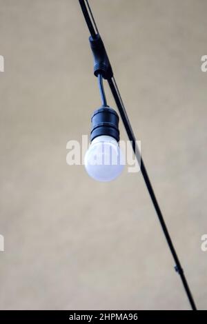 White light bulb is hanging on black wire. Part of a street garland on gray background. Close-up. Selective focus. Stock Photo