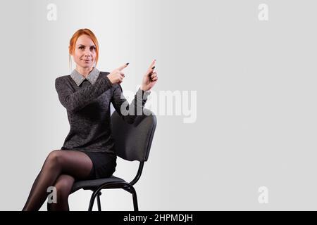 A girl, in business clothes, sits on a chair and indicates the direction with her hands, on a gray background Stock Photo