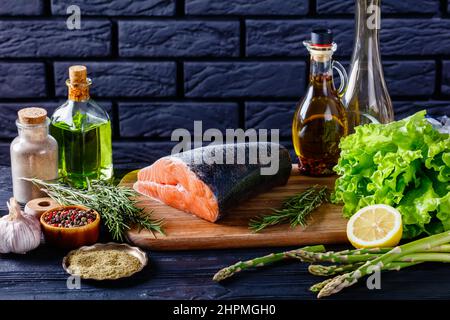 piece of raw salmon fish on a wood board with lettuce, rosemary, asparagus, lemon and spices with a black brick wall at the background Stock Photo