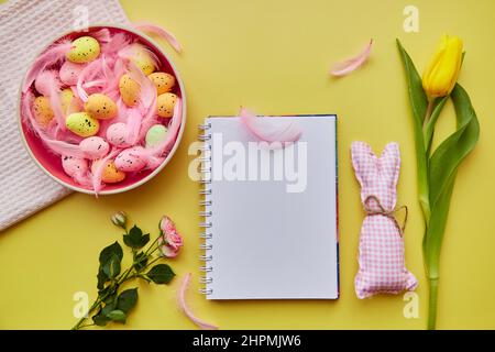 Easter mock up notepad. Pink bowl with colorful eggs, pink feathers, crafting cute bunny and flowers. Happy Easter concept. Top view. Post card mock up on yellow background Stock Photo