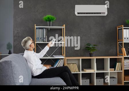 Man sitting on couch in his office switches modern air conditioner with remote control Stock Photo