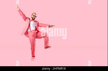 Cheerful young man in suit with suspenders makes funny movements on pastel pink background. Stock Photo