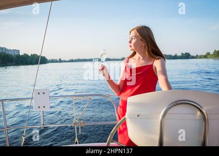 The elegant woman in a long red dress holding a glass of champagne standing on a yacht and looking for river sunset view Stock Photo