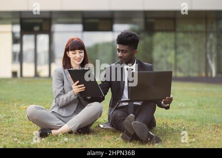 Caucasian woman in gray suit, holding clipboard while handsome African-American man signing contract. Two business colleagues in formal clothes sitting on grass outdoors in front of modern office Stock Photo