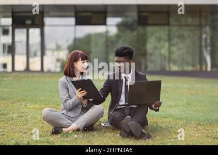 Caucasian woman in gray suit, holding clipboard while handsome African-American man signing contract. Two business colleagues in formal clothes sitting on grass outdoors in front of modern office Stock Photo