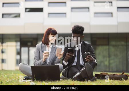 Coffee break. Businesspeople and technologies. Smiling happy African American businessman and his female Caucasian colleague, sitting on grass outdoors and using cellphone, drinking coffee Stock Photo