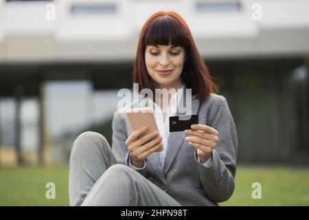 People, business, technologies, online payment. Beautiful smiling woman, sitting on grass outside, holds cellphone and credit card, makes shopping online or verifies account balance using mobile app Stock Photo