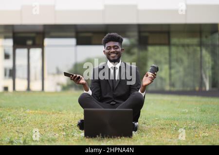 Office work and mindfullness. Handsome 30-aged African young business man meditating outside after work in front of company building, sitting on the grass with phone and coffee cup, with eyes closed Stock Photo