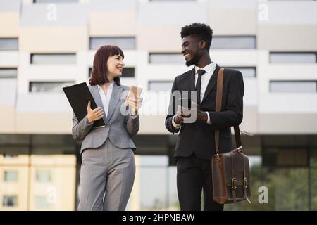 Front view of two happy smiling business colleagues, walking outside office and using smartphones. Pretty Caucasian woman and African American man in formal wear using phones outdoors Stock Photo