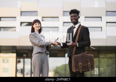 Cheerful smilin pretty Caucasian businesslady welcomes handsome African American male colleague, shaking hands near office building after successful job interview. Job hiring concept Stock Photo