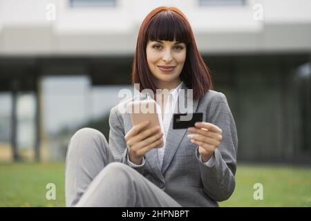 People, business, technologies, online payment. Beautiful smiling woman, sitting on grass outside, holds cellphone and credit card, makes shopping online or verifies account balance using mobile app Stock Photo