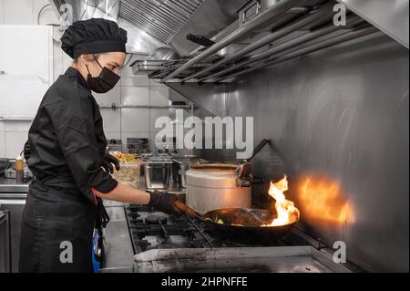 Woman Chef in protective face mask prepare food in the kitchen of a restaurant or hotel during new normal. Coronavirus prevention concept. High quality photo Stock Photo