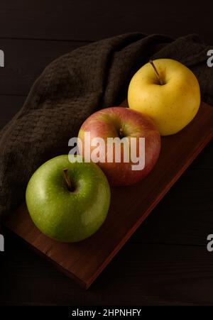 Three multicolored apples on a brown wooden board on a dark brown wooden background. Stock Photo