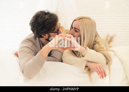 Cute Caucasian Couple Looking At Each Other Forming Heart Shape With Their Palms While Laying On Their Bed. High quality photo Stock Photo