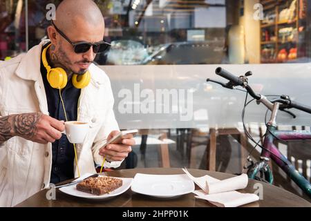 Latin young man with headphones drinks a cup of coffee while looking at his smart phone in a coffee shop next to his bicycle Stock Photo