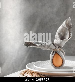 Festive Easter table setting. Funny Easter bunny made of egg and linen napkin on plate. Gray background. Stock Photo