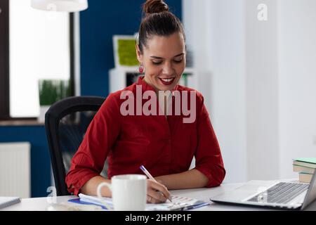 Entrepreneur writing notes on clipboard sitting at desk. Businesswoman with red shirt working in startup office in front of laptop. Smiling employee analyzing startup business chart. Stock Photo