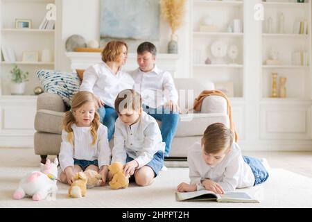 Happy caucasian parents are relaxing on cozy sofa in living room at home. Small children playing on a warm floor and reading a book. An overjoyed family rests on the weekend in their own home. Stock Photo