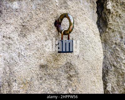 A small padlock hang on an iron hook, which is fixed in the wall, built of large blocks of limestone. Close-up. Selective focus. Copy space. Stock Photo