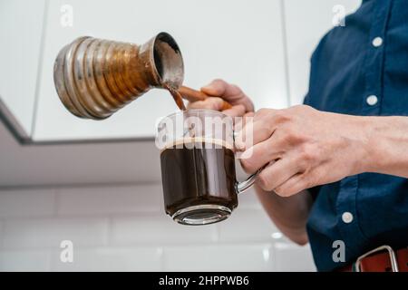 Close up of hands of unrecognizable man in blue shirt pouring fresh brewed hot natural coffee from copper cezve with wooden handle into glass cup in w Stock Photo