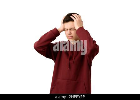 Handsome teenage boy suffering from headache or migraine, isolated on white background. Young guy wearing maroon hoodie wincing in pain Stock Photo