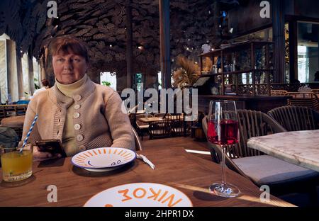 A pretty adult mature woman sits at a restaurant table with a gadget in her hand and a glass of wine and orange fresh juice on the table. Stock Photo