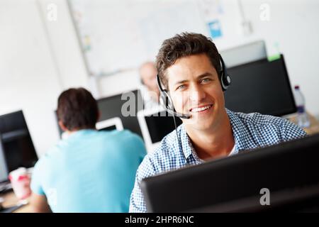 Hes happy to be of service. Portrait of a handsome young sales assistant talking on his headset while working on a computer and colleagues working in Stock Photo