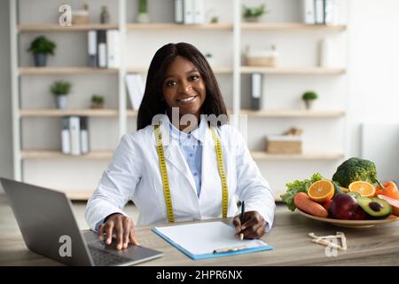 Cheerful african american female dietologist working with laptop, having fresh fruits and vegetables on table Stock Photo