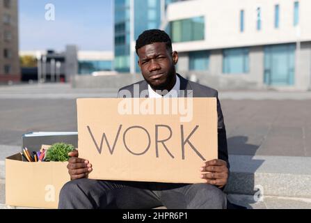 Portrait of fired black businessman with paper poster saying work and box of personal things, sitting on stairs outdoors Stock Photo