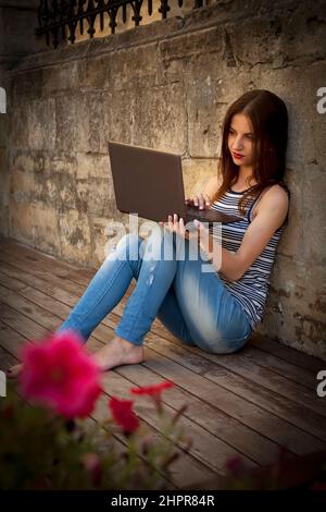 Beautiful young business woman freelance typing on laptop outdoors on a wooden floor. Best places to work remotely in a city. Vertical image. Stock Photo