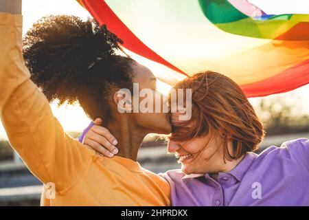 Happy women gay couple having tender moment holding rainbow flag outdoor - Main focus on african girl eye Stock Photo