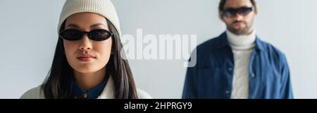 asian woman in white beanie and dark sunglasses looking at camera isolated on grey, banner Stock Photo