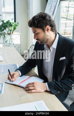 stylish businessman writing in empty notebook near papers on office desk Stock Photo