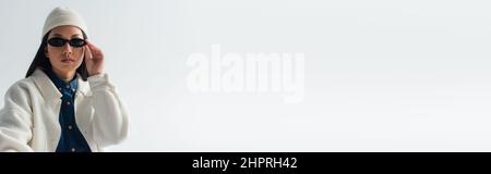 fashionable asian woman in white jacket and beanie adjusting sunglasses isolated on grey, banner Stock Photo