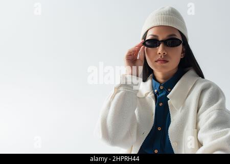 young asian woman in white jacket and beanie adjusting dark sunglasses isolated on grey Stock Photo