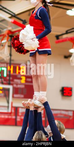 A high school cheerleader is standing tall high in the air with her teammates holding her up wearing facemasks while cheering. Stock Photo